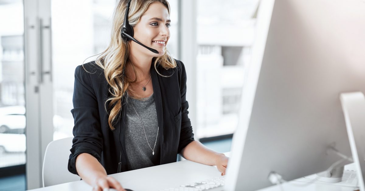 Shot of a young woman using a headset and computer at her desk in a modern office